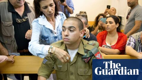 A man in military uniform sits on a wooden bench in a courtroom and is comforted by a woman.