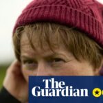 Monica Dolan, as Catherine Corless, touches her ear with a concerned expression outdoors